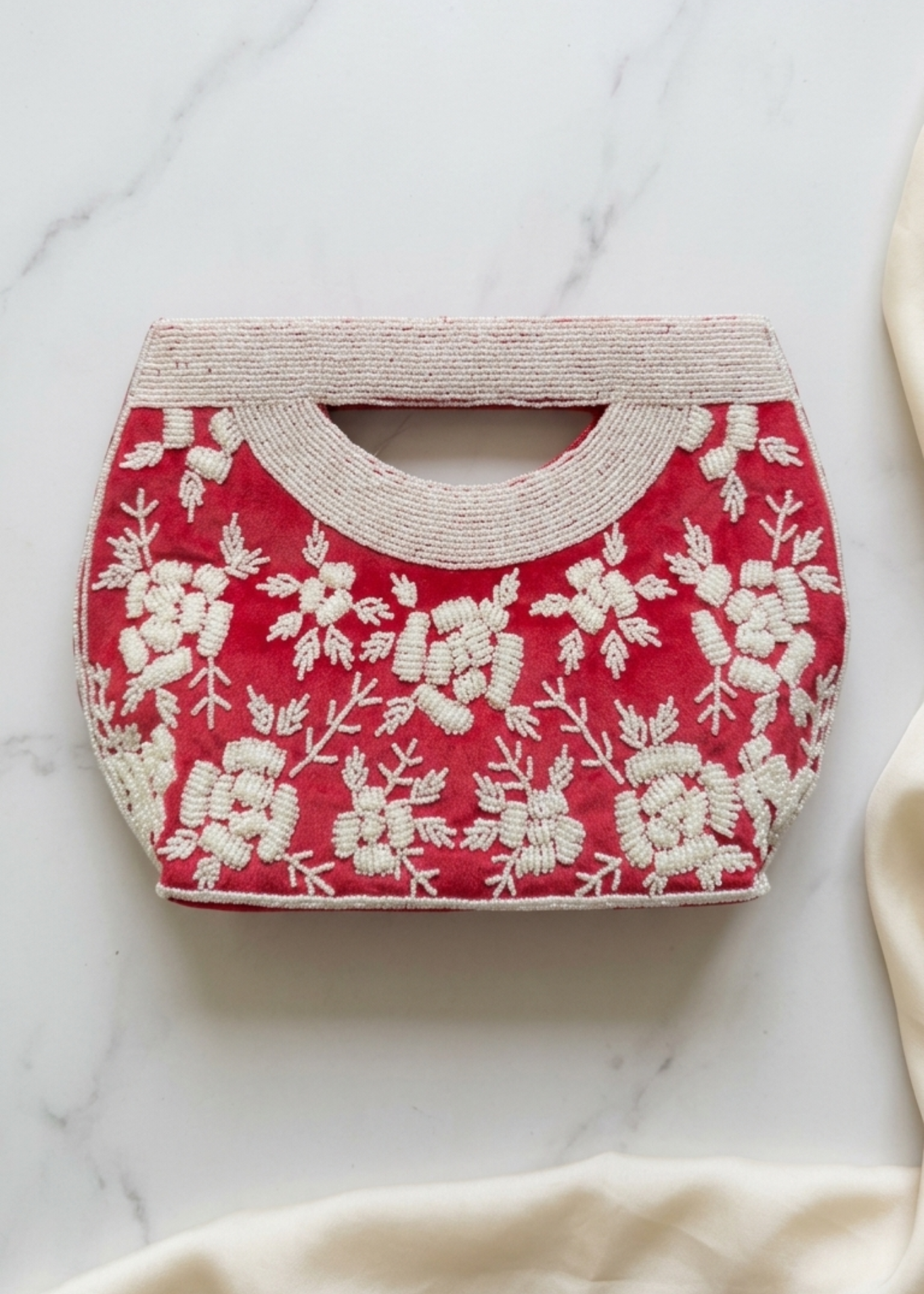 A red velvet clutch bag with intricate white floral embroidery, featuring a curved top handle and resting on a white marble surface with a soft cream fabric draped in the foreground.